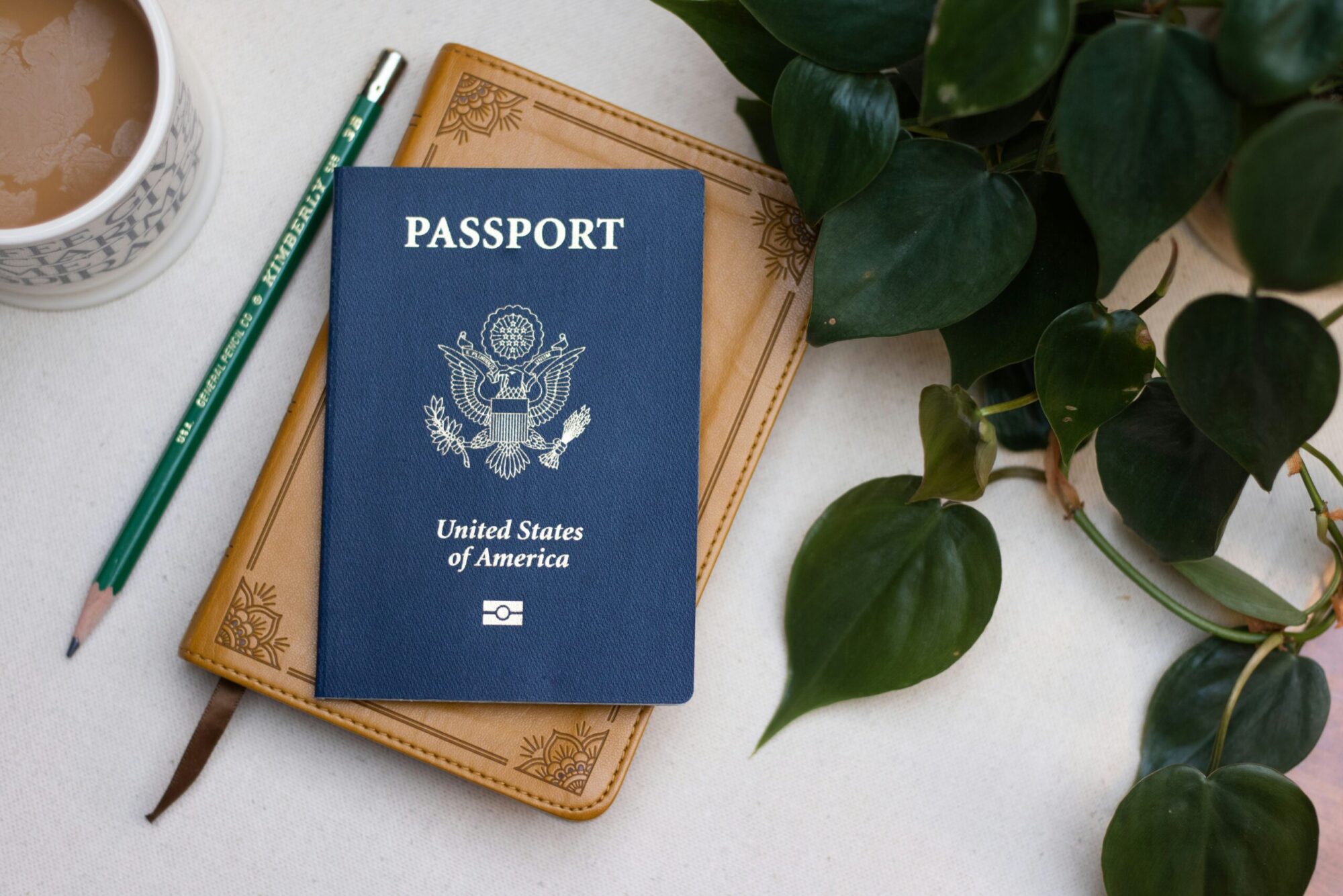 Picture of a US passport on a table surrounded by a cup of coffee, a pencil and a plant